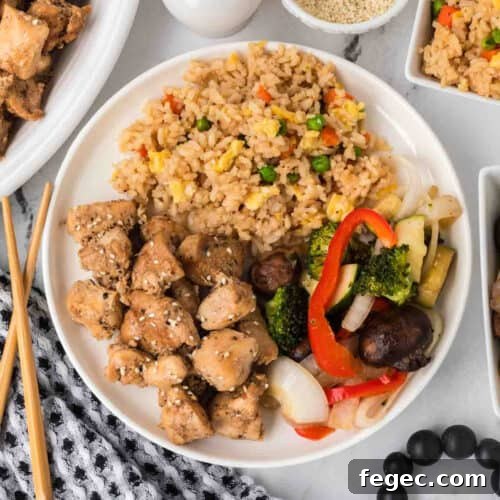 A plate of Hibachi Chicken with sesame seeds, fried rice with peas and carrots, and sautéed vegetables.