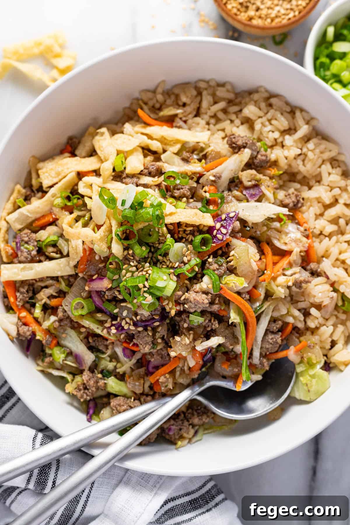 Bowl of rice topped with egg roll in a bowl ground beef mixture, wonton strips, green onions, and sesame seeds.