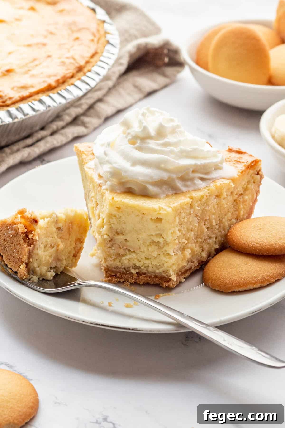 slice of banana pudding cheesecake topped with whipped cream and served on a white round plate with two vanilla wafers on the side and a forkful of the cheesecake