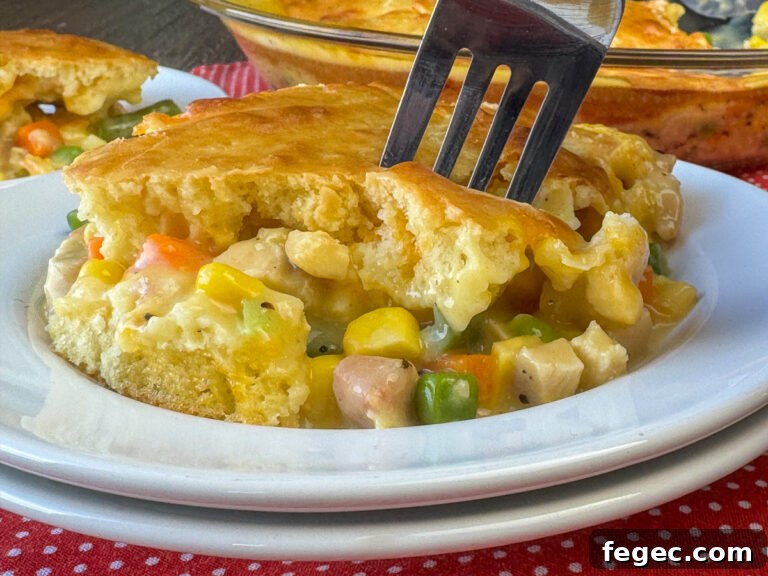 Close up fork in a top layer of fluffy Bisquick baked over a creamy chicken and vegetable filling on a white plate with the full pot pie in the background.