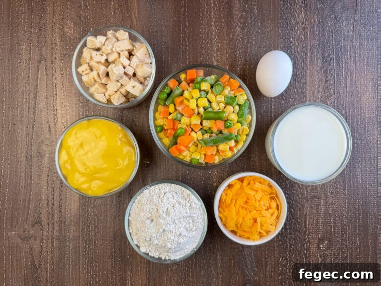 Ingredients to make an easy chicken pot pie are condensed cream of chicken soup, mixed vegetables, cubed chicken, an egg, milk, cheddar cheese and flour on a wooden counter top.