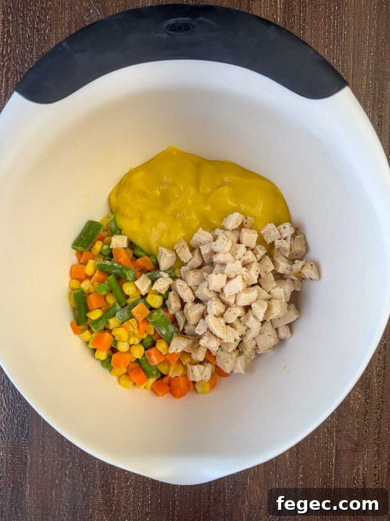 Can of condensed cream of chicken soup, corn, carrots, peas, green beans and small cubes of chicken poured into a white mixing bowl.