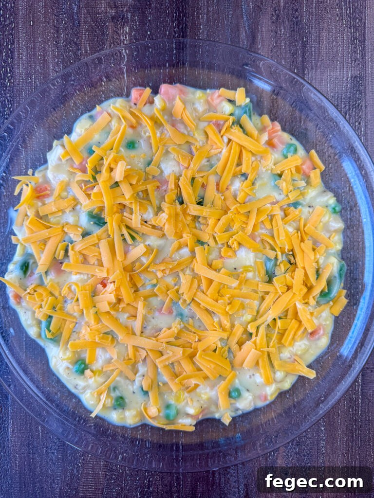 Cheddar cheese sprinkled over a chicken soup and vegetable mixture in a glass pie plate on a wooden counter top.