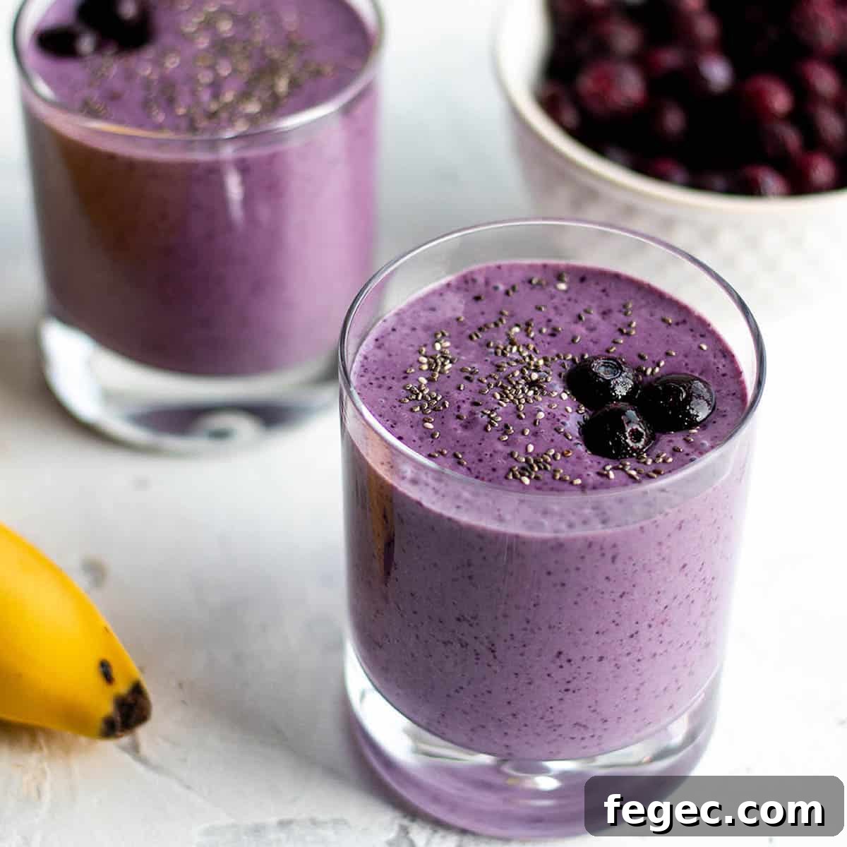 Two small glasses with a purple smoothie topped with blueberries with a bowl of blueberry in the back and a banana.