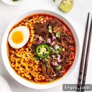 Bowl of beef birria ramen with soft boiled eggs, lime wedges, chopped onions and cilantro.